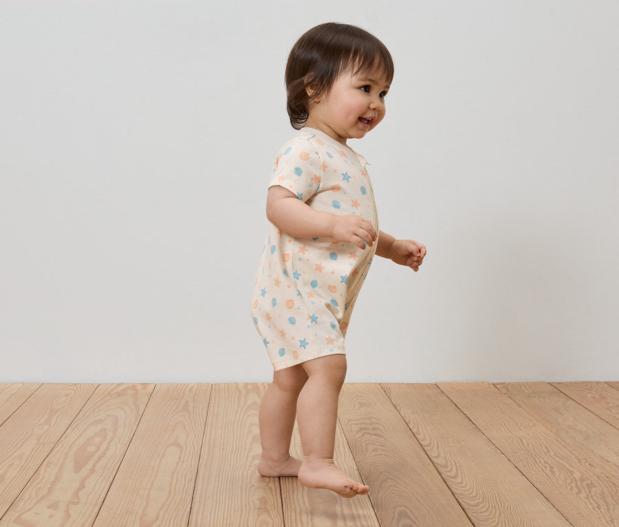 Ein Kleinkind mit braunen Haaren trägt einen hellen Spielanzug mit blauem und orangefarbenem Muster und geht auf einem Holzboden.