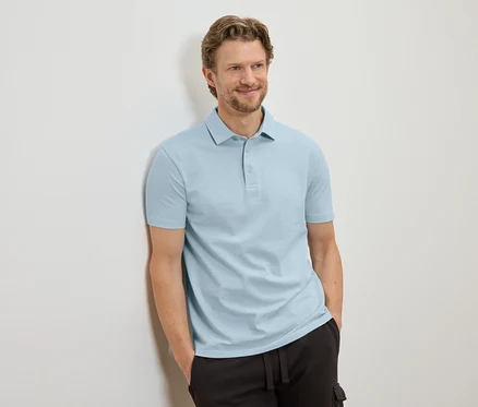 Mann mit hellbraunen Haaren, der an einer Wand lehnt und ein hellblaues Poloshirt und eine dunkle Hose trägt.