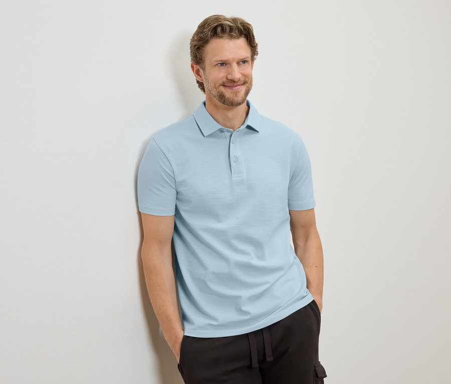 Mann mit hellbraunen Haaren, der an einer Wand lehnt und ein hellblaues Poloshirt und eine dunkle Hose trägt.