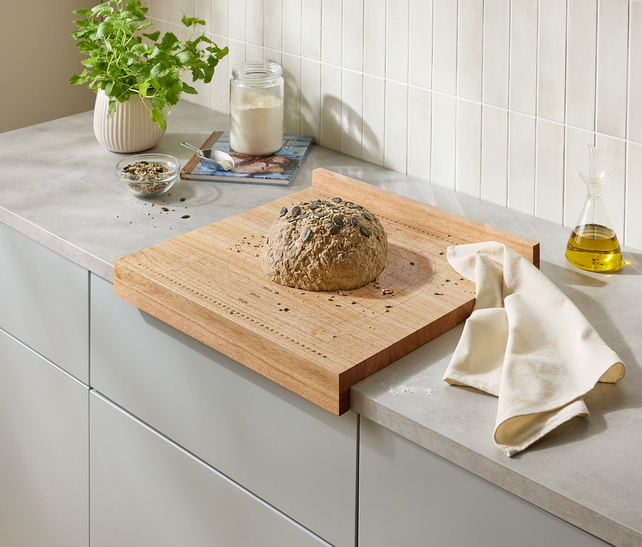Ein Laib Brot mit Kürbiskernen liegt auf einem Holzbrett auf einer Küchentheke. Neben dem Brot liegt ein beiges Tuch.