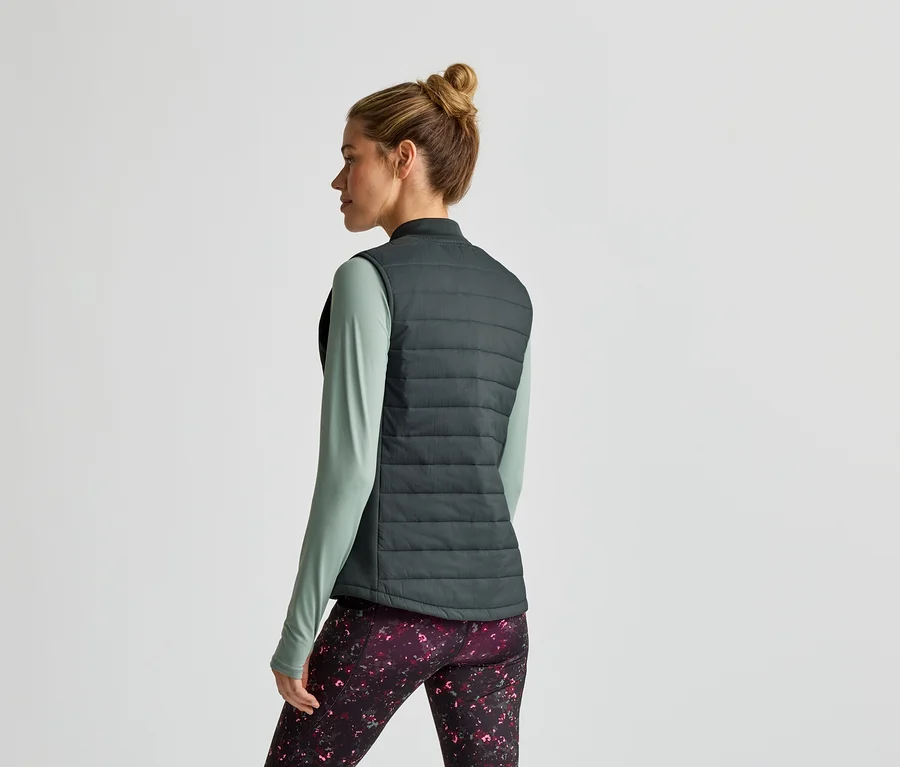 Frau von hinten in grüner Weste, Shirt und Leggings mit Muster vor hellem Hintergrund.