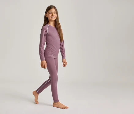 Ein lächelndes Mädchen mit langen braunen Haaren geht barfuß und trägt ein lila Langarmshirt und passende Leggings mit hellen Streifen.