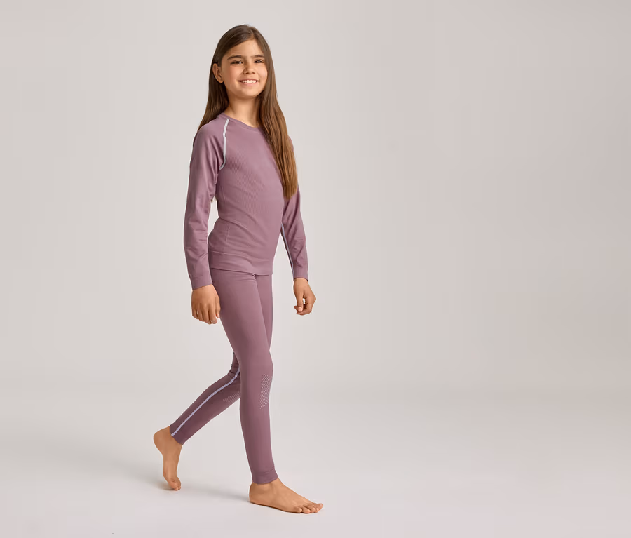 Ein lächelndes Mädchen mit langen braunen Haaren geht barfuß und trägt ein lila Langarmshirt und passende Leggings mit hellen Streifen.
