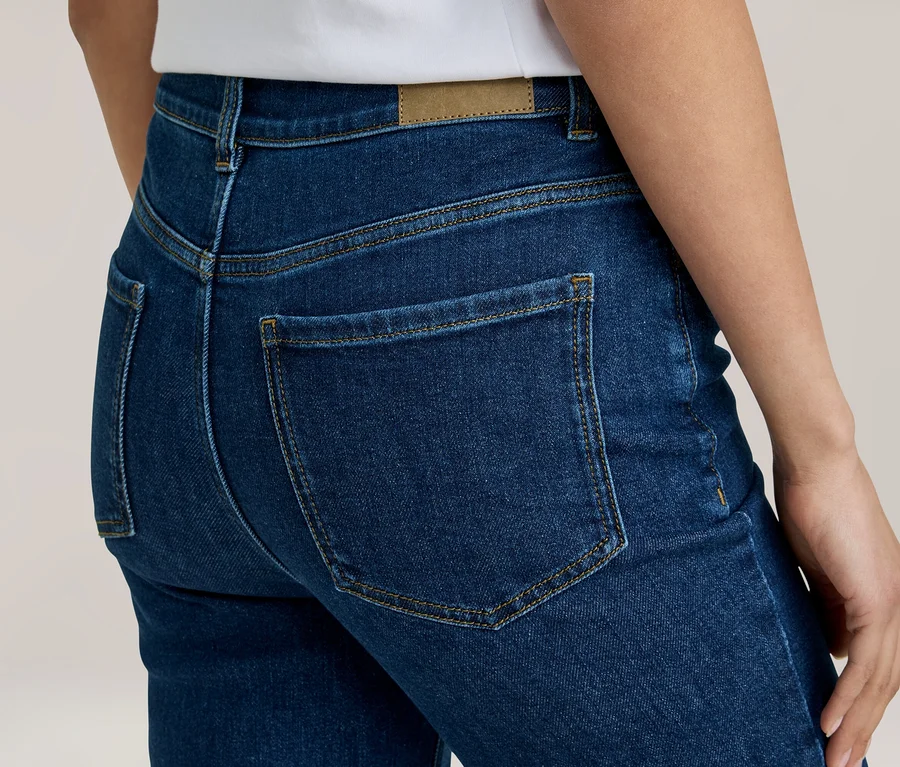 Rückenansicht einer Person in dunkelblauen Jeans mit sichtbaren Gesäßtaschen, gelben Nähten und einem braunen Etikett am Bund. Ein weißes Shirt ist oben sichtbar.