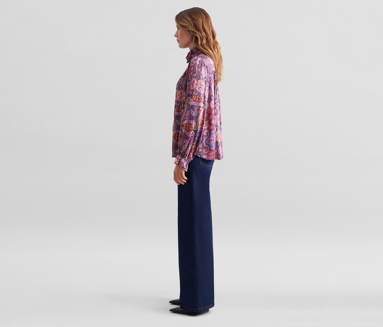 Frau im Profil mit einer lila-pinken Paisley-Bluse mit Rüschenkragen und dunklen weiten Jeans.