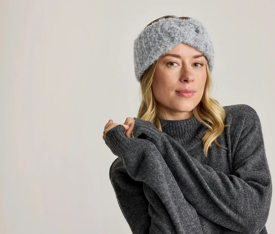 Eine blonde Frau trägt ein graues Strickstirnband und einen grauen Pullover, blickt nach vorne und hat die Hände verschränkt.