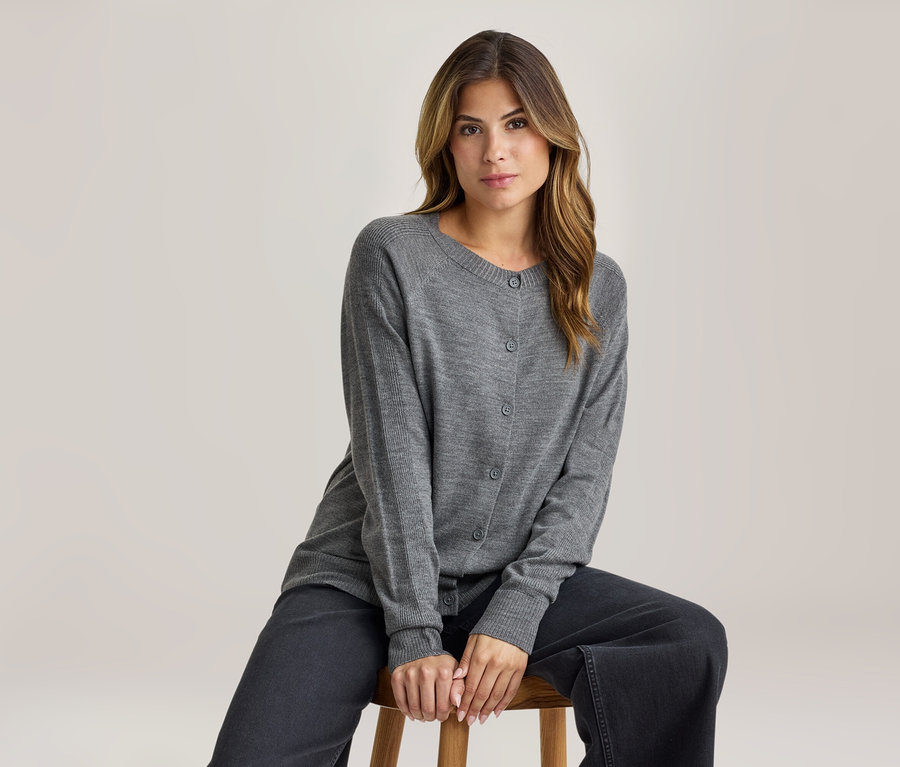 Frau sitzt auf einem Holzschemel, trägt einen grauen Strickcardigan und eine dunkelgraue weite Hose, blickt nach vorne.