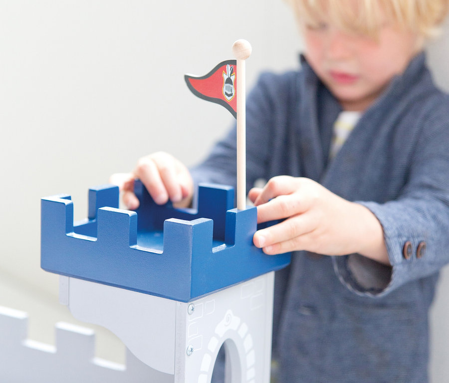 Ein kleiner Junge mit blonden Haaren spielt mit einer grau-blauen Holzspielzeugburg und richtet den blauen oberen Teil aus. Eine rote Flagge steckt auf der Burg, eine kleine Puppe liegt darin.