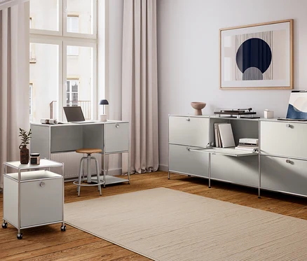 Ein modernes Homeoffice mit hellgrauen modularen Möbeln. Ein Schreibtisch mit Laptop und schwarzer Lampe steht vor einem großen Fenster. Unter dem Schreibtisch befindet sich ein Hocker mit Holzsitzfläche. Links daneben steht ein mobiler Rollcontainer mit Pflanze und Kerze. Rechts ist ein langes Aufbewahrungsmöbel mit offenem Fach, Büchern, Plattenspieler und Kunstwerk. Ein großer Teppich liegt auf dem Holzboden.