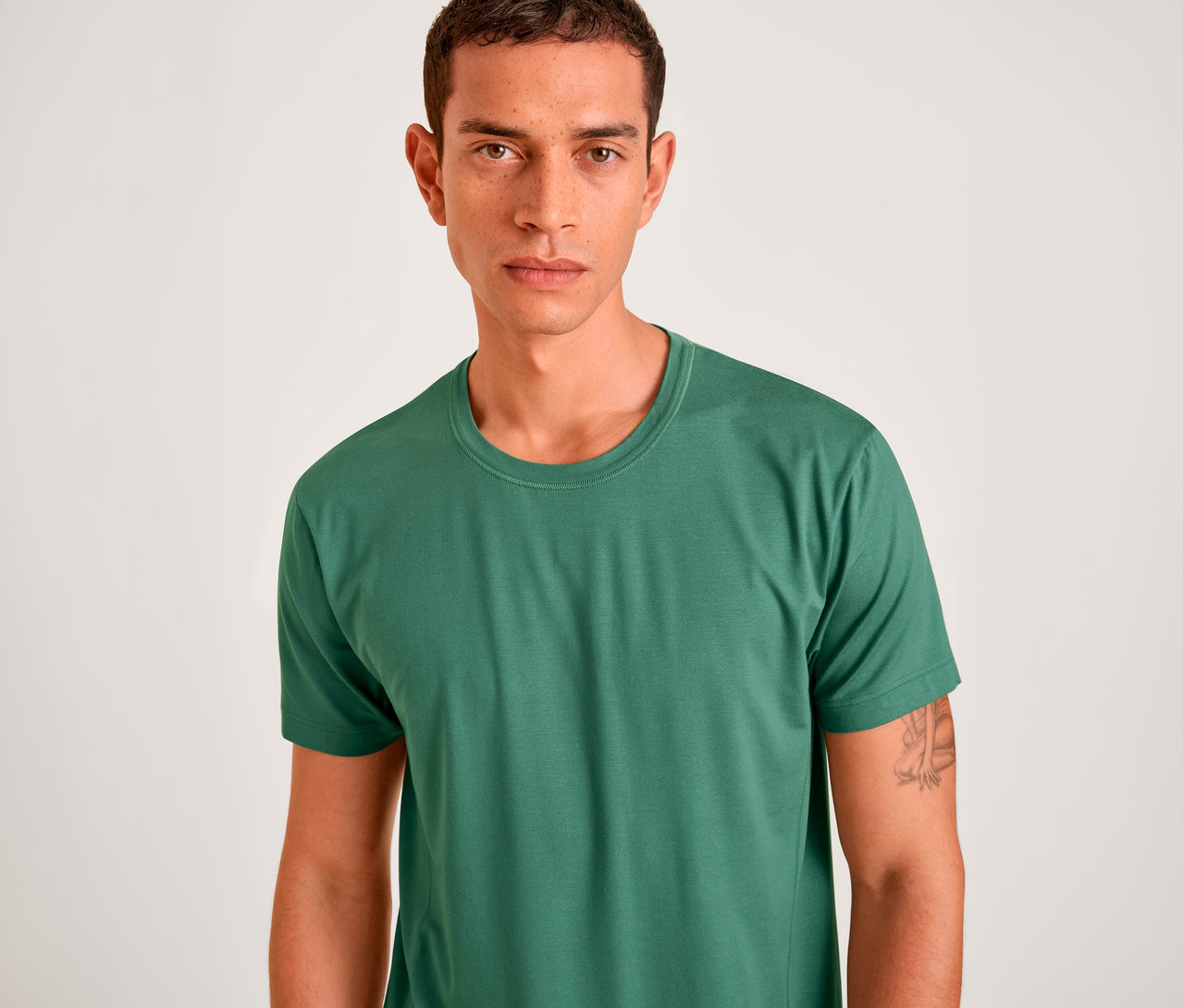 Mann mit kurzen dunklen Haaren und Sommersprossen trägt ein grünes, kurzärmeliges T-Shirt mit Rundhalsausschnitt und blickt direkt in die Kamera. Ein Tattoo ist auf seinem rechten Arm sichtbar.