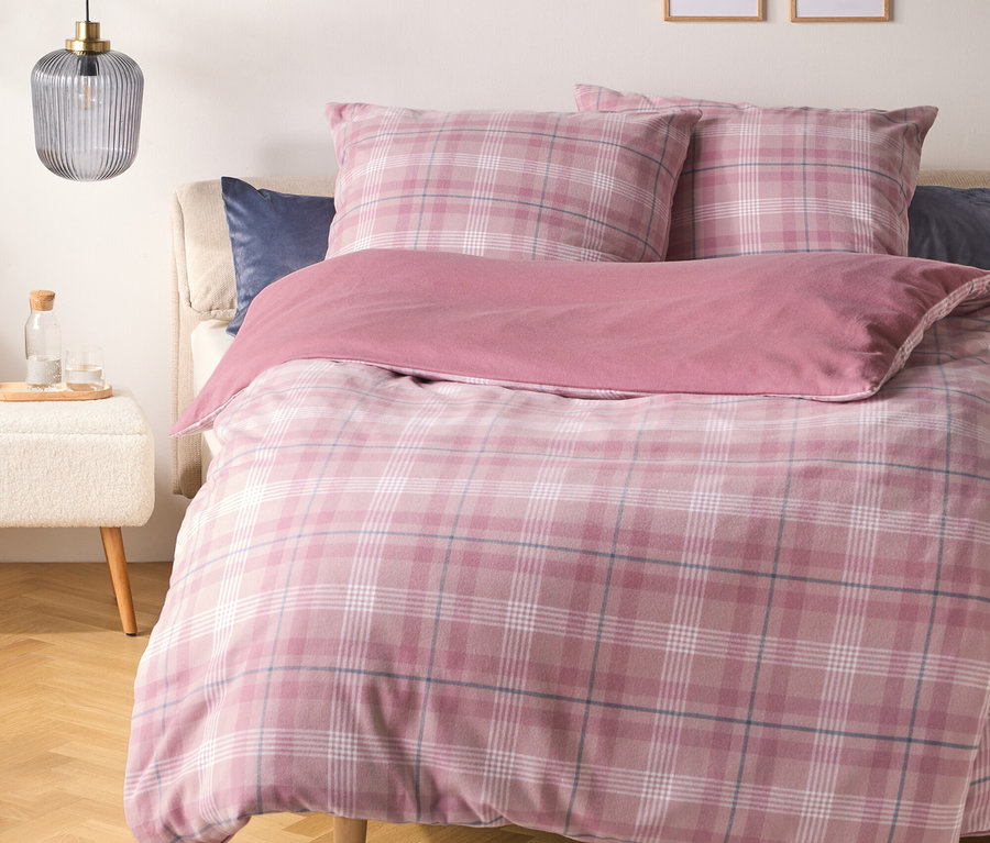 Ein Bett mit rosa karierter Bettdecke und Kissen sowie einer einfarbigen rosa Decke. Ein dunkelblaues Kissen liegt hinter den karierten Kissen. Links steht ein weißer Hocker mit einer Glasflasche und zwei Gläsern unter einer gerippten grauen Pendelleuchte. Zwei gerahmte Strichzeichnungen hängen an der Wand über dem Bett.