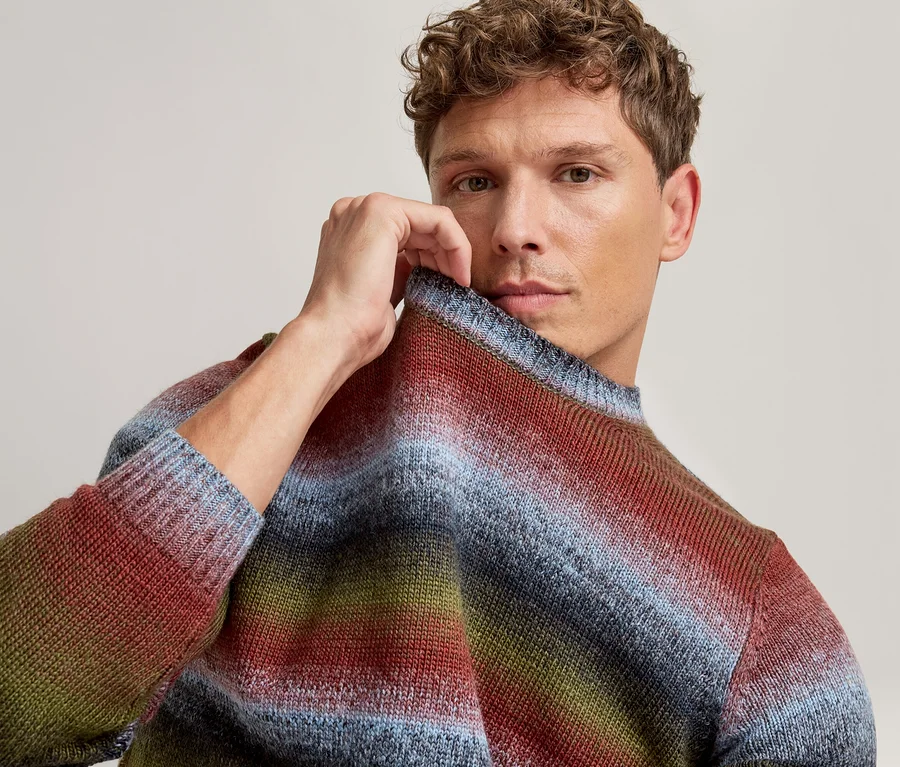 Mann mit lockigem Haar, der den Kragen seines blau, rot, grün und braun gestreiften Pullovers hält und in die Kamera blickt.