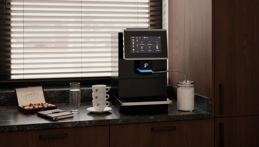 Schwarze Kaffeemaschine mit Touchscreen und blauem Licht auf einer Küchenarbeitsplatte. Daneben stehen ein Milchbehälter, gestapelte weiße Tassen mit Tchibo-Logo, ein Wasserglas, eine offene Lindt Pralinenschachtel und ein braunes Notizbuch. Im Hintergrund sind Holzschränke und ein Fenster mit Jalousien zu sehen.