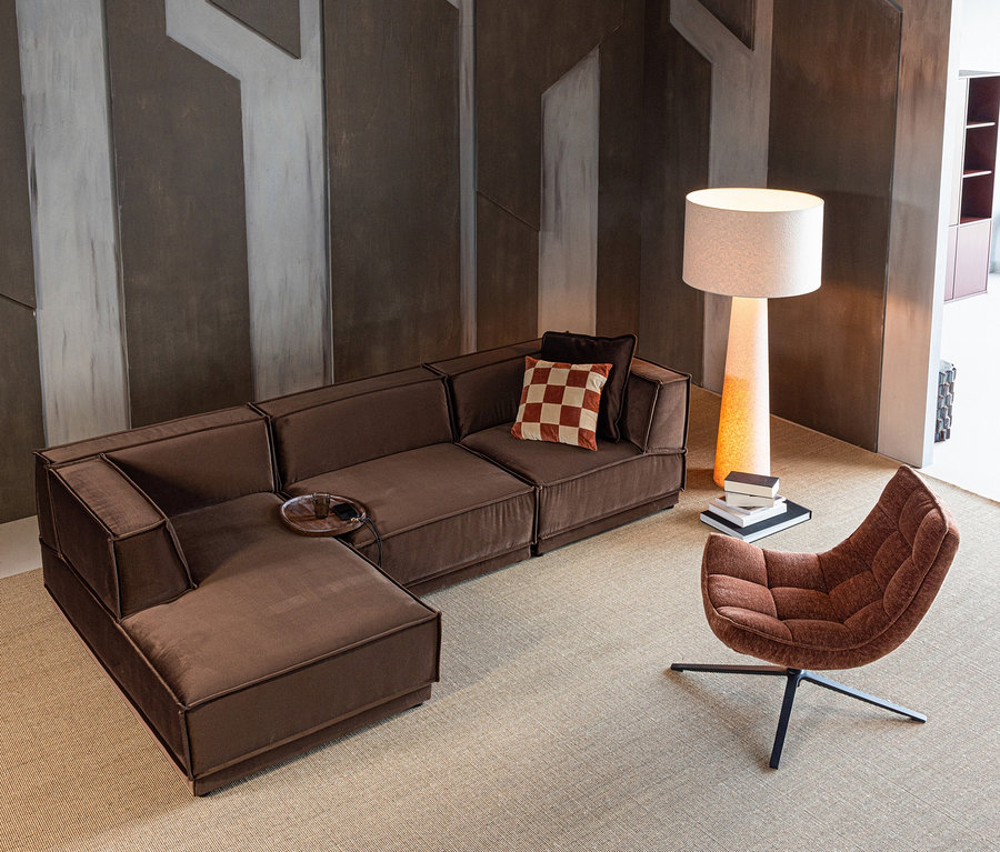Braunes L-Sofa, brauner WOOOD Drehsessel »Maudi«, Stehlampe und Bücher auf hellem Teppich vor geometrischer Wand.