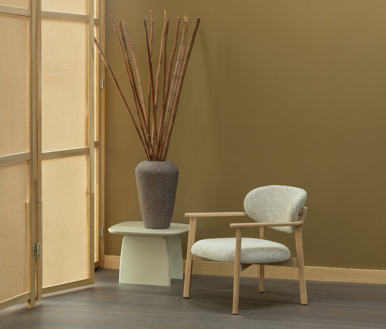 Ein moderner Raum mit senfgelber Wand und dunklem Boden. Links steht ein heller Raumteiler aus Holz. Ein heller Beistelltisch trägt eine hohe, strukturierte graue Vase mit trockenen Ästen. Rechts davon steht ein Sessel mit hellem Holzrahmen und hellgrauem Polster.