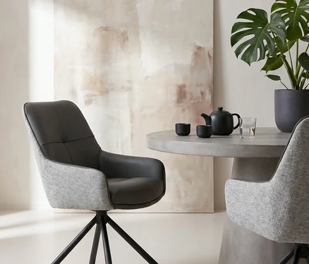 Zwei moderne Stühle mit dunkelgrauem und hellgrauem Stoff stehen um einen runden Betontisch mit einer schwarzen Teekanne, zwei Tassen und einem Glas Wasser. Rechts steht eine große Monstera-Pflanze in einem dunklen Topf, im Hintergrund ein abstraktes Gemälde in Beigetönen.