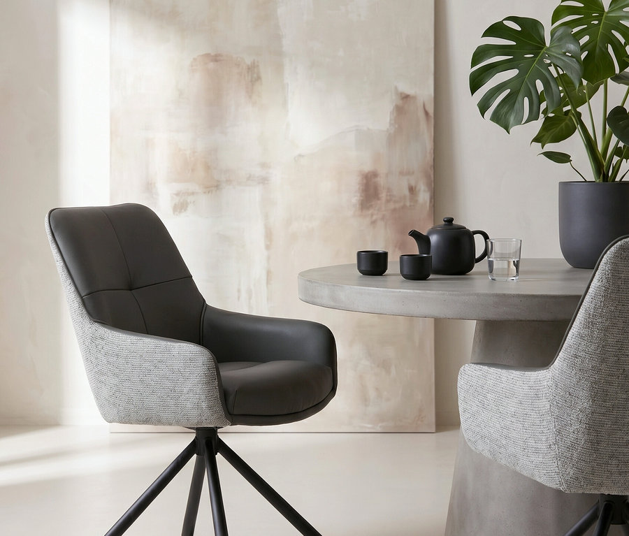 Zwei moderne Stühle mit dunkelgrauem und hellgrauem Stoff stehen um einen runden Betontisch mit einer schwarzen Teekanne, zwei Tassen und einem Glas Wasser. Rechts steht eine große Monstera-Pflanze in einem dunklen Topf, im Hintergrund ein abstraktes Gemälde in Beigetönen.