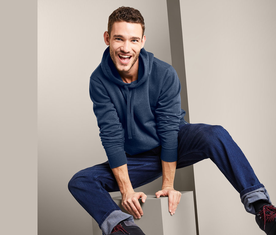 Lächelnder Mann sitzt auf einem grauen Würfel, trägt einen blauen Hoodie und Jeans.