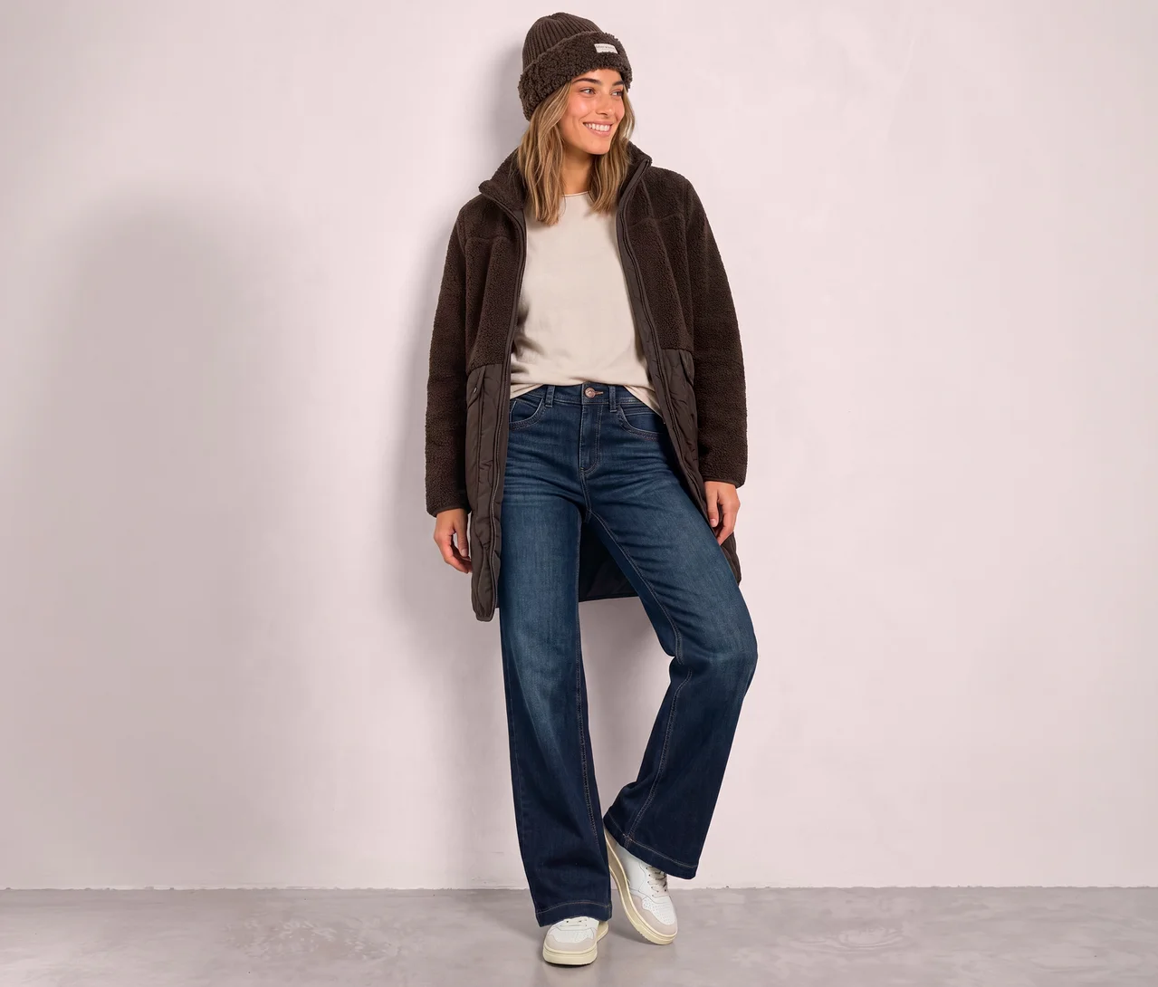 Lächelnde Frau in einem Street One Teddymantel, beigem Oberteil, blauen Jeans und einer braunen Mütze.