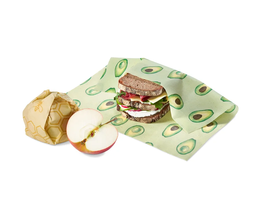 Ein Sandwich, eingewickelt in ein Gaia-Bienenwachstuch mit Avocados, daneben ein Apfel und ein eingewickelter Gegenstand.