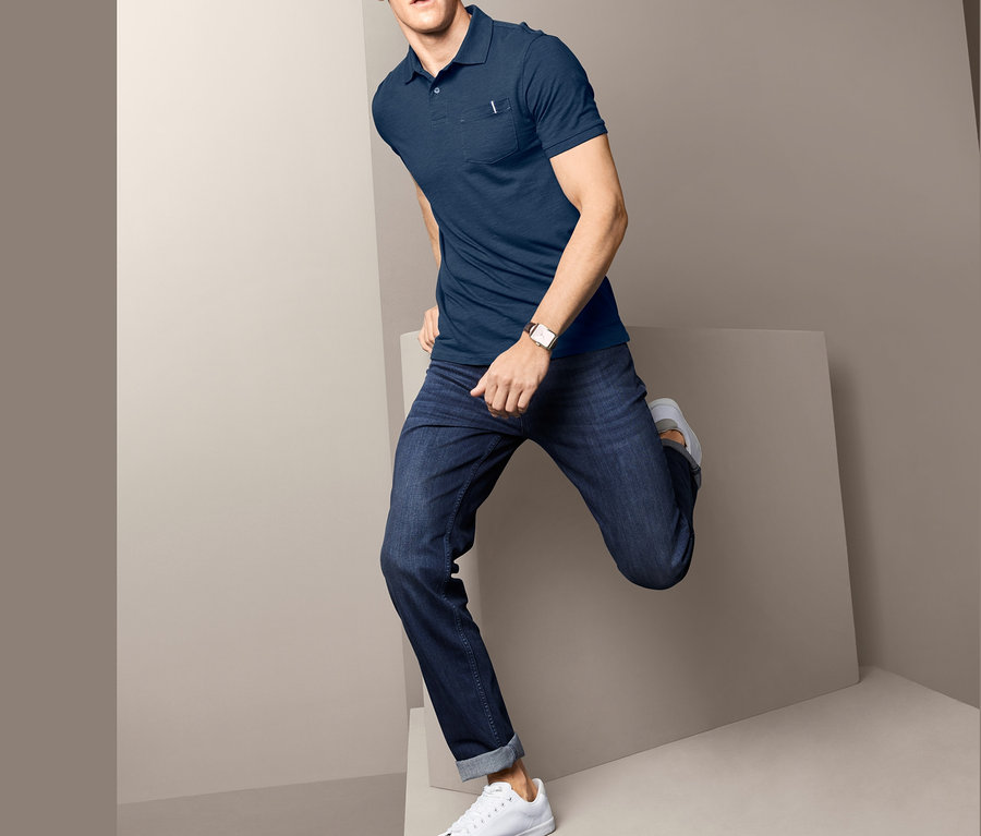 Mann posiert in blauem Poloshirt mit Strukturgarn, Jeans und weißen Schuhen vor grauem Hintergrund.
