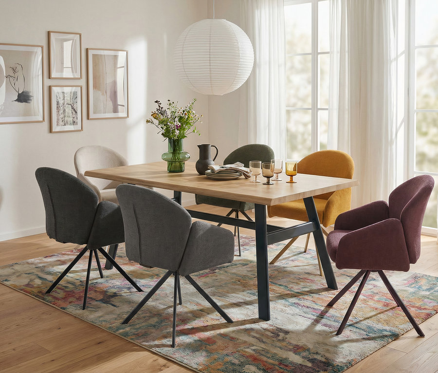 Ein modernes Esszimmer mit hellem Holztisch und sechs bunten Polsterstühlen auf einem gemusterten Teppich. Auf dem Tisch stehen eine Blumenvase, ein Krug, Teller und Gläser. Eine Papierlaterne hängt darüber, mit gerahmter Kunst an der Wand und transparenten Vorhängen.