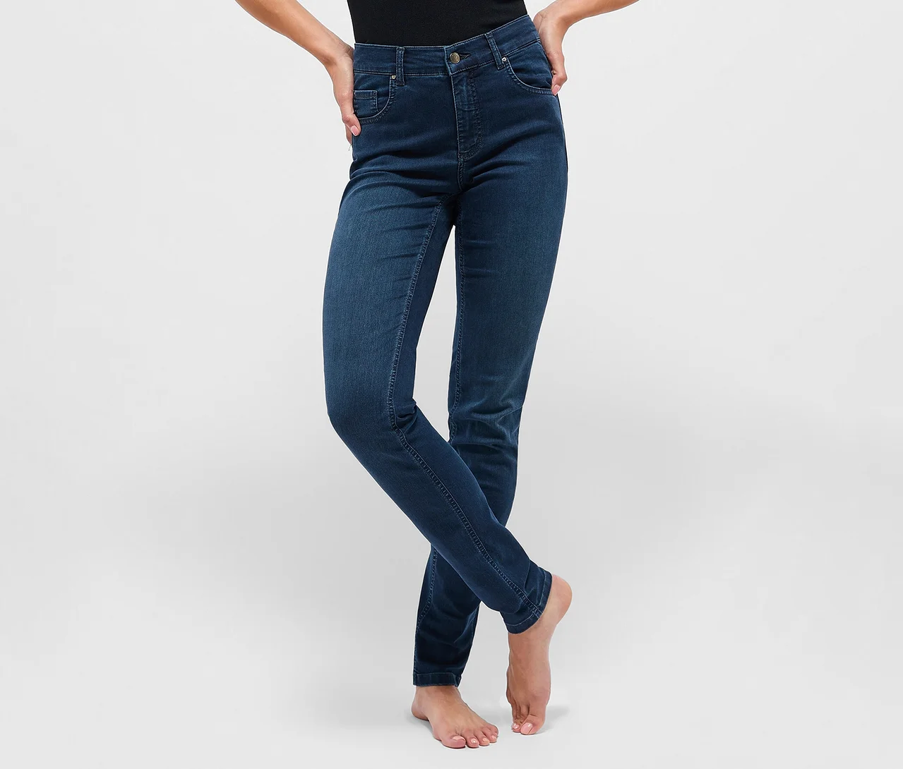 Person in dunkelblauer, hochgeschnittener Slim-Fit-Jeans, barfuß stehend mit Händen in den Hüften.
