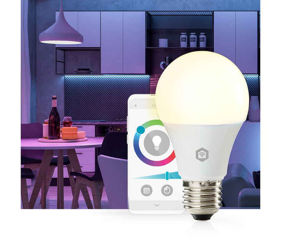Eine smarte Glühbirne, die warmweißes Licht ausstrahlt, steht vor einem Smartphone, das eine Lichtsteuerungs-App mit Farbrad und Helligkeitsregler anzeigt. Im Hintergrund ist eine moderne Küche und ein Essbereich mit lila und blauem Umgebungslicht zu sehen.