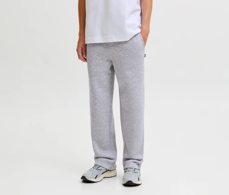 Person trägt graue Jack&Jones Bradley Sweatpants, ein weißes T-Shirt und graue Turnschuhe, eine Hand in der Hosentasche.