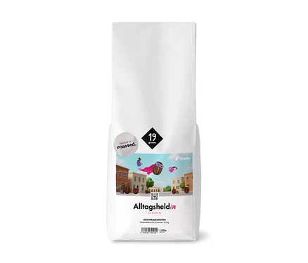 Weißer, aufrecht stehender Kaffeebeutel mit der Aufschrift "Alltagsheldin" und "Tchibo". Er zeigt eine Illustration eines fliegenden Kaffeehelden mit pinkem Umhang über einer Stadt und die Geschmacksnoten "Amarenakirsche, Brownie, Honig" sowie "1000g".