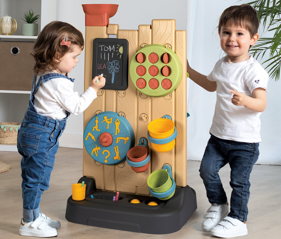 Zwei Kinder spielen mit einer Smoby Life Activity Wall. Ein Mädchen in Jeans-Latzhose schreibt mit Kreide auf einer Tafel, während ein Junge daneben steht und lächelt.