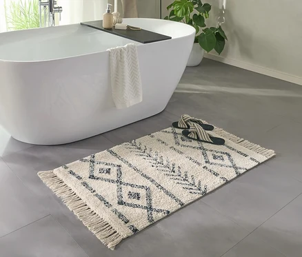 Cremefarbene benuta Badematte "Naomi" mit dunklen Mustern und Fransen auf grauem Fliesenboden vor einer weissen Badewanne mit Handtuch und Badeschlappen.