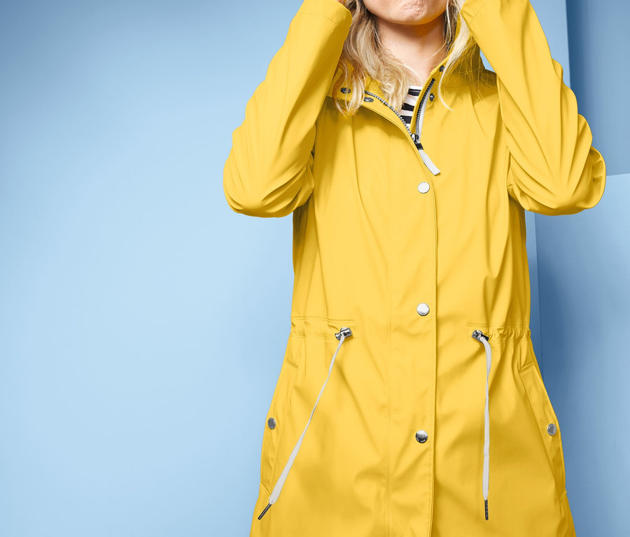 Frau zieht Kapuze vom gelben Regen-Parka über die Augen und spitzt die Lippen.