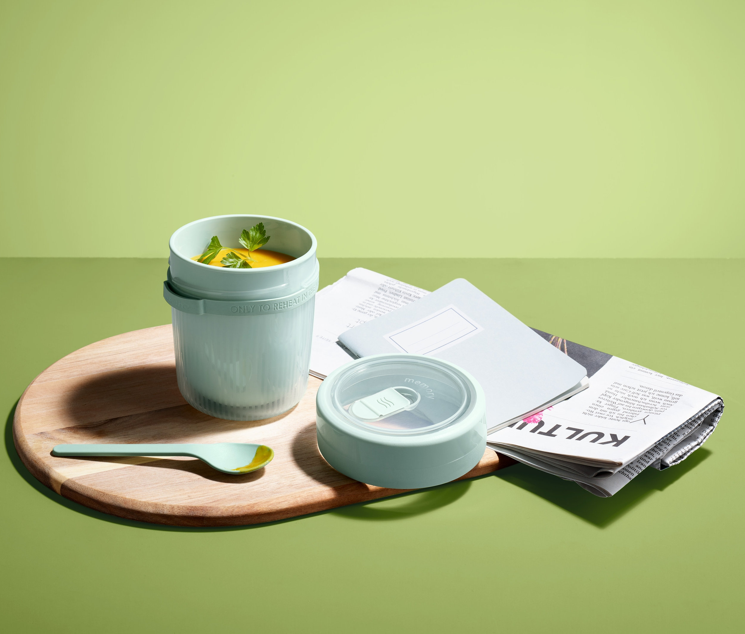 To-go-Suppenbehälter mit Löffel auf einem Holzbrett neben einer Zeitung und einem Notizblock.