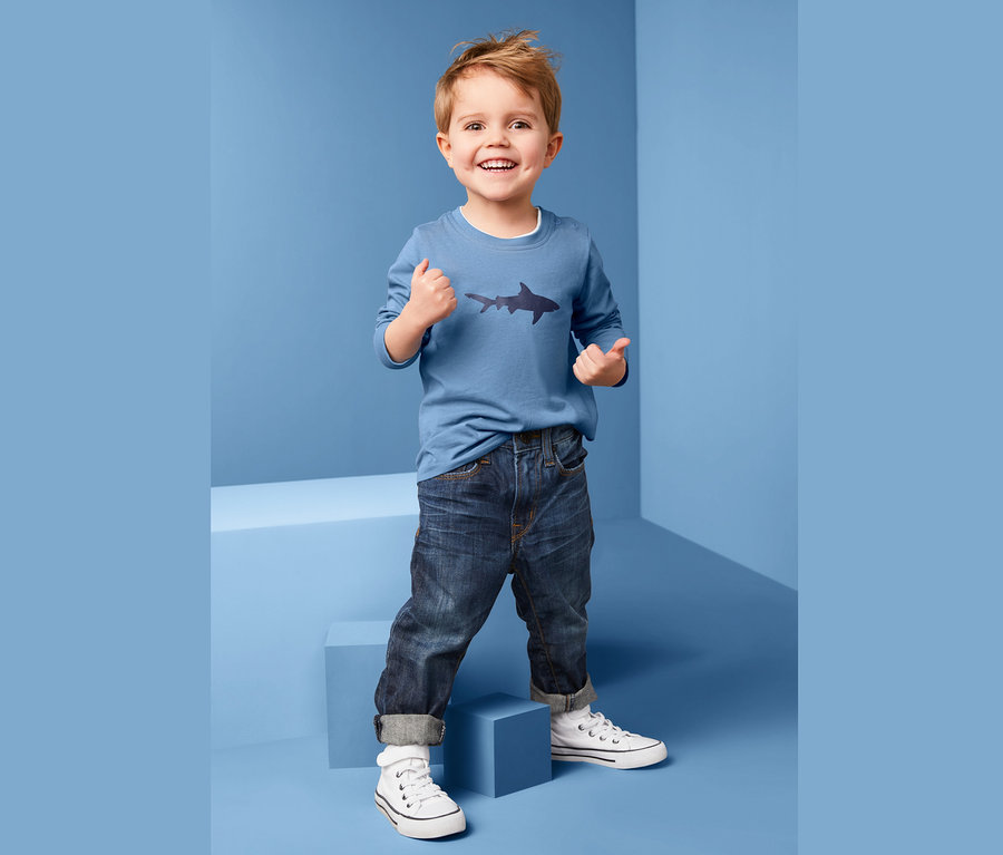 Ein Junge in einem blauen Langarmshirt mit Hai-Motiv lächelt und zeigt die Daumen nach oben.