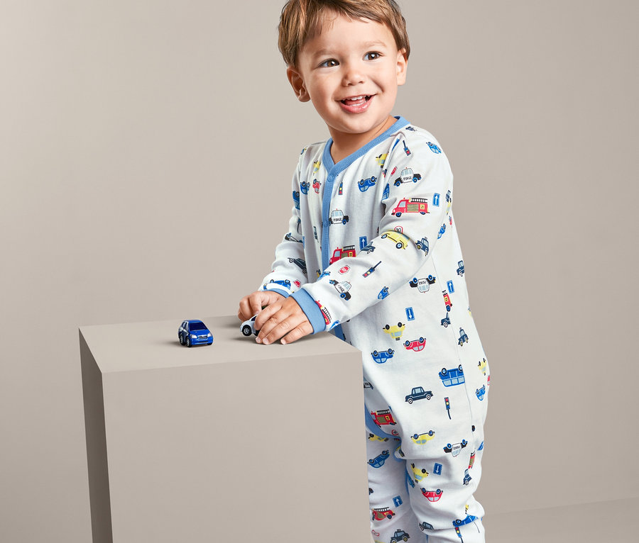 Ein kleiner Junge steht und spielt mit Spielzeugautos auf einem grauen Würfel. Er trägt einen 2 Pyjamas mit Automotiv.