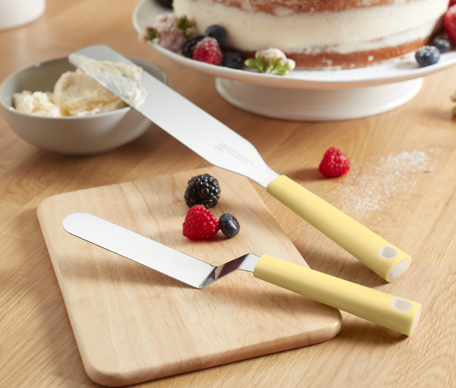 Zwei Streichpaletten liegen auf einem Holzbrett, im Hintergrund ein mit Früchten dekorierter Kuchen.