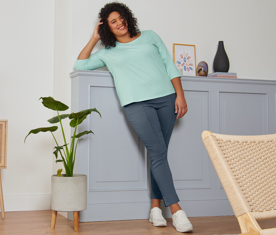 Eine Frau lehnt an einer Wand und lächelt. Sie trägt ein mintfarbenes Shirt mit 3/4-Arm, eine rauchblaue Stretchhose und Veloursleder-Sneaker.