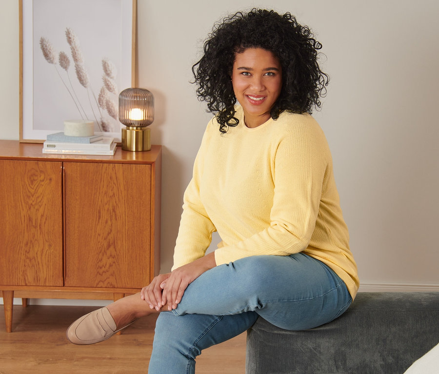 Frau sitzt auf einem grauen Sofa und trägt einen gelben Strickpullover, Treggings und Loafer aus Veloursleder.
