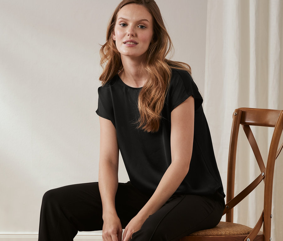 Frau sitzt auf einem Stuhl und trägt eine schwarze Hose und ein Shirt mit Satinfront.