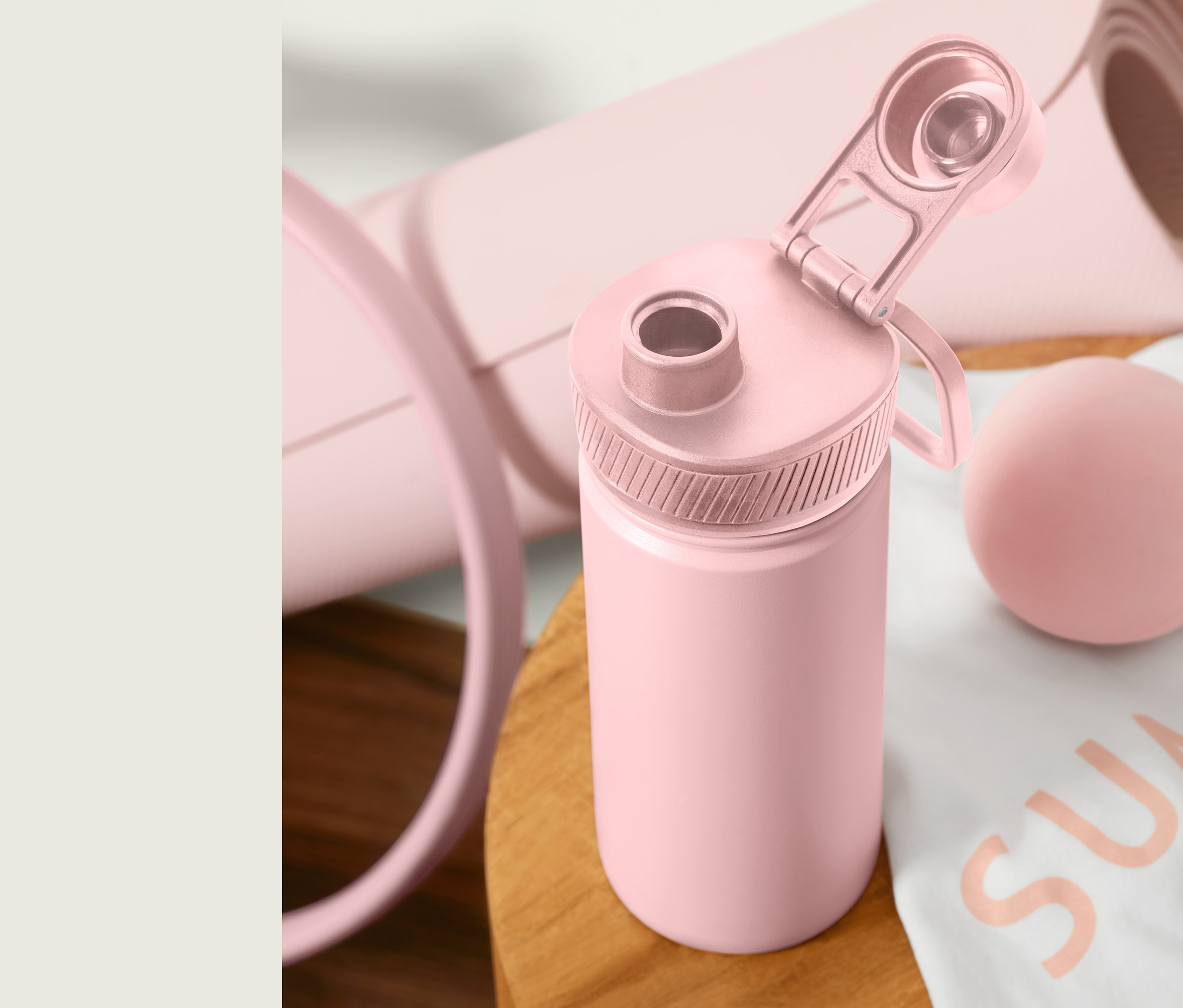 Eine rosa Sport-Trinkflasche mit geöffnetem Deckel steht auf einem Holztisch neben einer zusammengerollten Yogamatte und einem Massageball.