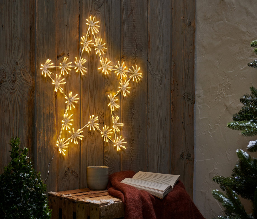 Ein LED-Outdoor-Stern »Cluster« hängt an einer Holzwand. Auf einer Holzkiste liegen ein offenes Buch und eine Tasse.