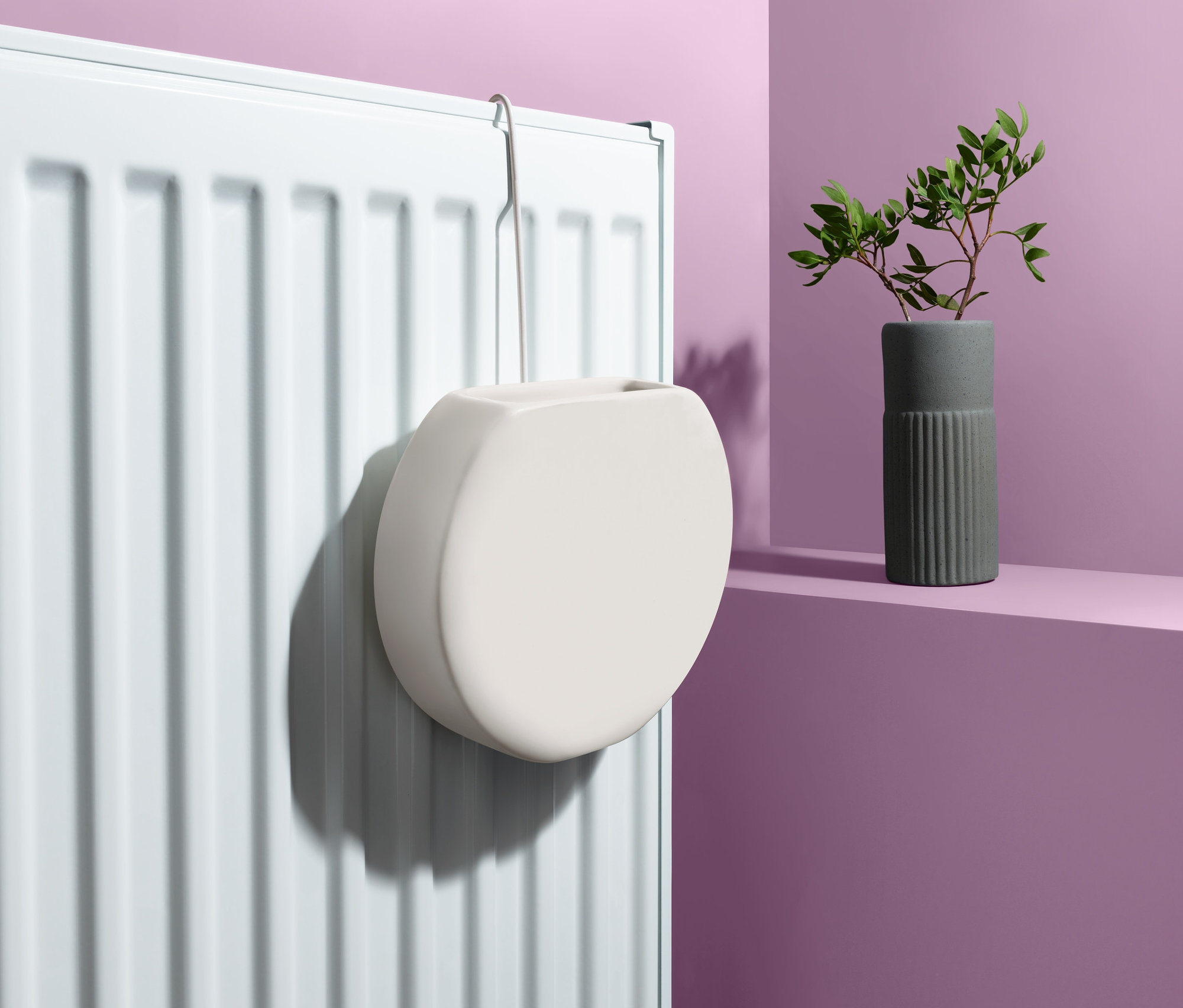 Ein weißer Heizkörper-Luftbefeuchter hängt an einem weißen Heizkörper. Daneben steht eine Vase mit grünen Blättern.