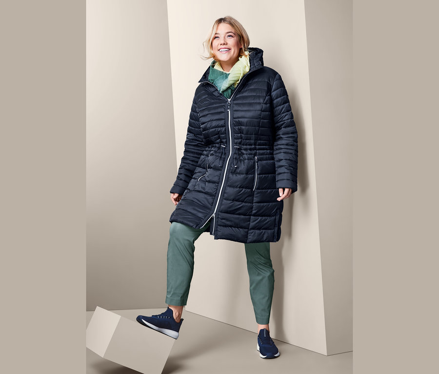 Eine Frau steht auf einem Würfel und trägt eine knöchellange Stretchhose und einen Steppmantel mit Kapuze.