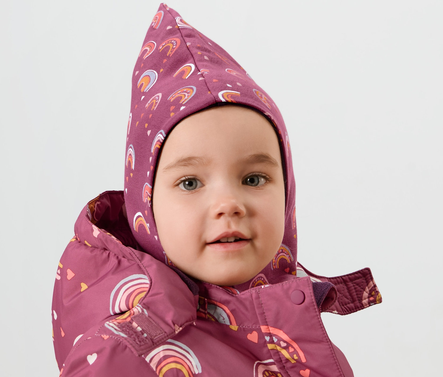 Porträt eines Kindes, das eine Kinder-Wende-Schlupfmütze mit Regenbogenmuster und eine Jacke mit Regenbogenmuster trägt.