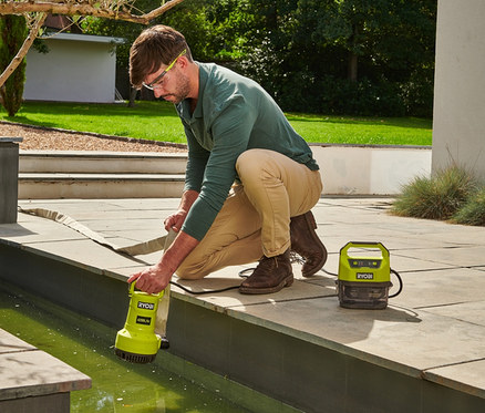 Mann mit Schutzbrille kniet an einem grünen Wasserbecken und hält eine RYOBI 18 V One+ Akku-Tauchpumpe ohne Akku und Ladegerät.