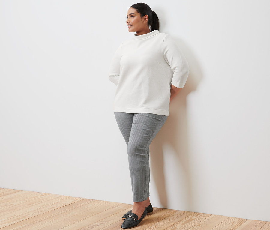 Eine Frau lehnt an einer Wand und trägt ein cremefarbenes Sweatshirt mit 3/4-Arm, eine Stretchhose und Leder-Loafer.