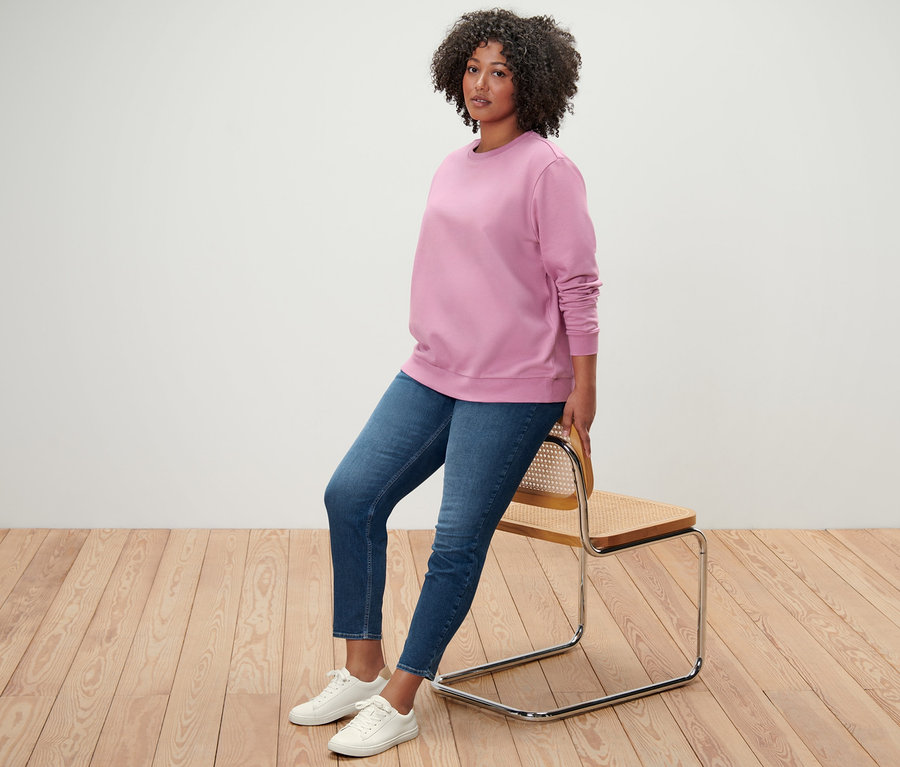 Eine Frau sitzt an einen Stuhl gelehnt und trägt ein rosa Sweatshirt, Treggings »Fit Nelly« und weiße Turnschuhe.