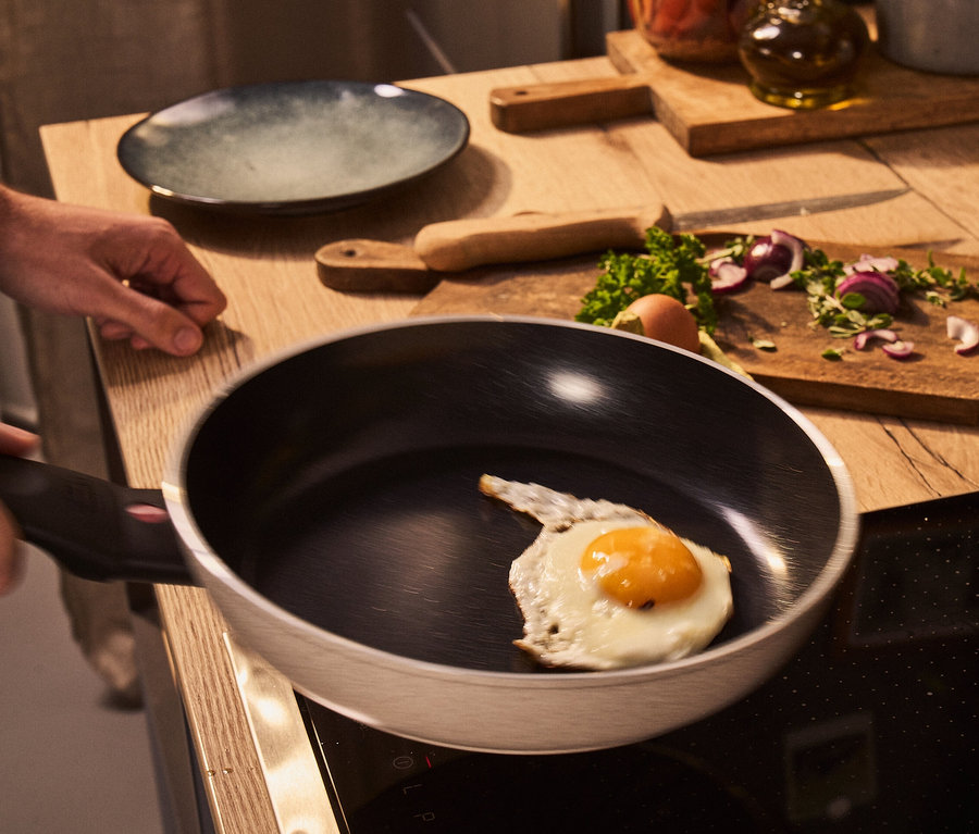 Mann brät ein Ei in einer BALLARINI Aluminium-Bratpfanne, sand, 28 cm. Auf der Küchentheke liegen geschnittenes Gemüse und andere Küchenutensilien.