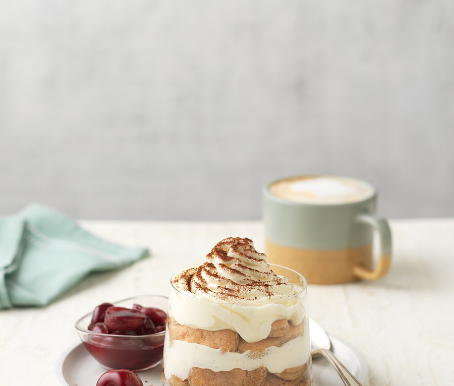 Dessert im Glas mit Kirschen, Sahne und Kakao auf einem Teller neben einem Löffel, einer Kaffeetasse und einem blauen Tuch.